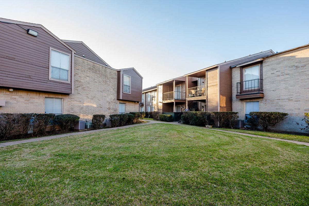 View of a residential apartment complex featuring two buildings with a landscaped courtyard. The buildings have a mix of brick and wood siding, and there are balconies visible. Neatly trimmed grass and hedges create an inviting outdoor space between the structures.