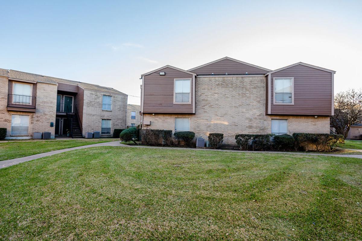 Two-story apartment buildings with brown accents are surrounded by well-maintained green lawns. The first building has multiple windows and a staircase leading to the entrance, while the second building features unique rooftop extensions. The background shows a clear sky, indicating a bright day.