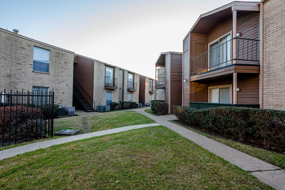 A view of a residential apartment complex featuring two buildings with balconies, green lawns, and a pathway. One building has stairs leading to upper units. The scene is set during the evening with soft lighting.
