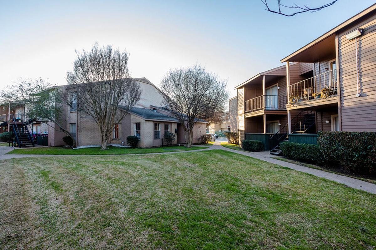 A view of a residential area featuring two multi-unit buildings, surrounded by neatly trimmed grass and a few trees. One building is a brick structure, while the other has a wooden exterior with balconies. The scene is well-lit, suggesting a pleasant day.