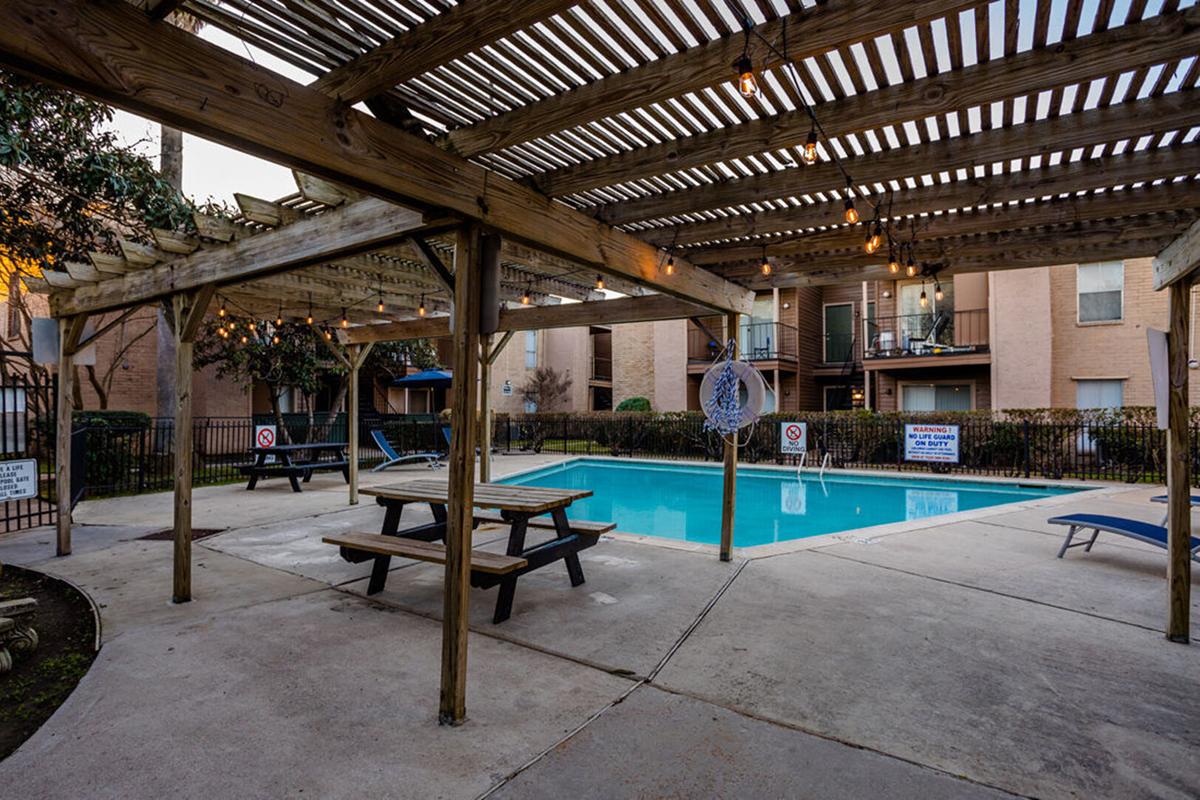 A swimming pool area with a wooden pergola overhead, surrounded by greenery and an apartment building. The pool is clear and inviting, with lounge chairs and a picnic table nearby. String lights hang from the pergola, creating a cozy atmosphere. Signs indicate pool rules.
