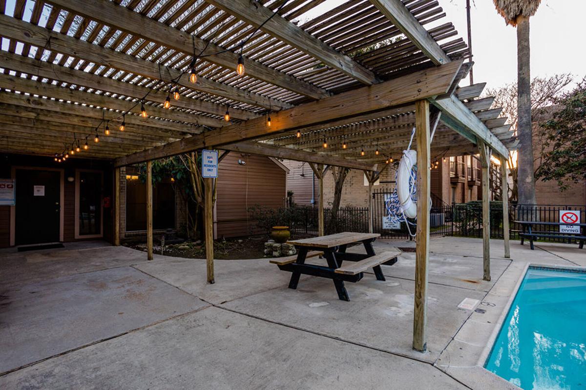 A covered patio area with a picnic table and hanging lights, adjacent to a swimming pool. The patio features wooden beams and is surrounded by a fence. In the background, there are apartment buildings and decorative plants.