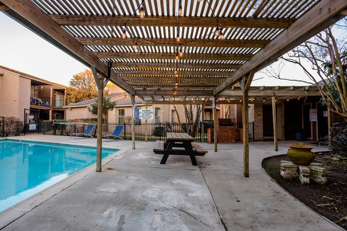 A pool area featuring a wooden pergola with string lights, picnic table, and lounge chairs. Surrounding the pool is a concrete patio, with apartment buildings visible in the background, creating a cozy outdoor setting for relaxation and socializing.