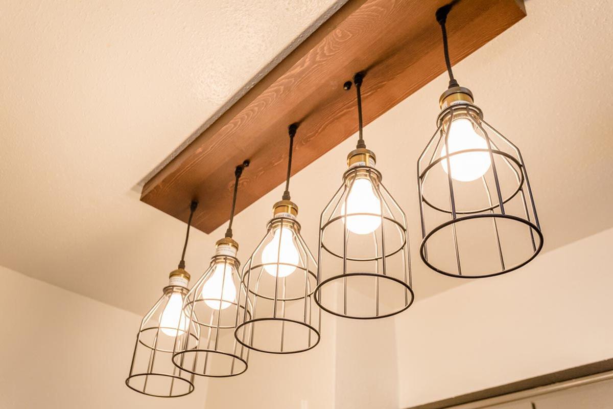 A rustic lighting fixture featuring five exposed light bulbs enclosed in wire cage designs, suspended from a wooden panel on the ceiling. The overall look combines modern and industrial styles, providing a warm, inviting illumination for the space.