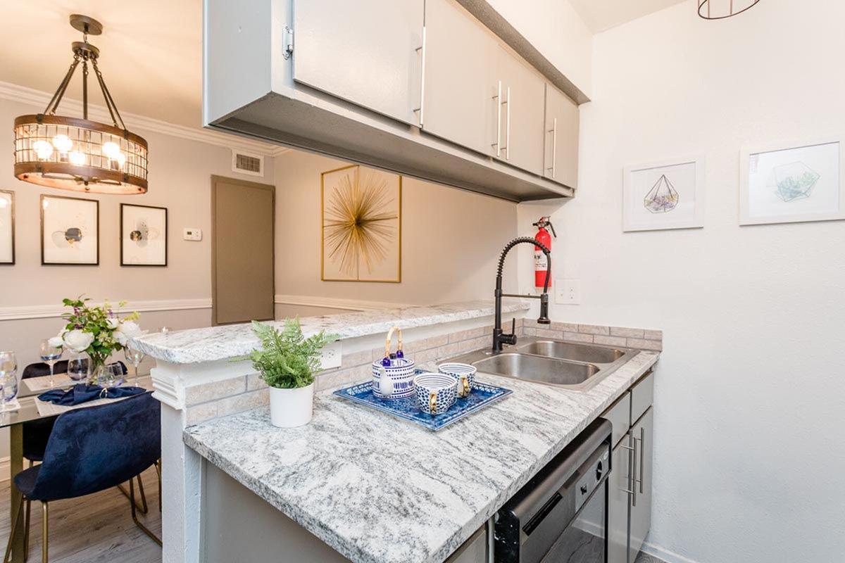 A modern kitchen featuring a granite countertop, stainless steel sink, and dishwasher, with a decorative tray holding dishes. In the background, there are framed artworks and a dining area with a dark table set for dining, alongside a wall-mounted fire extinguisher and a stylish chandelier.