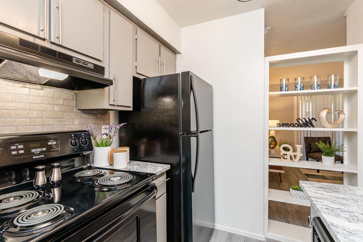 A modern kitchen featuring a black stove and refrigerator, light-colored cabinets, and a decorative shelf with plants and a welcoming sign. The countertop is made of a marble-like material, creating a clean and inviting atmosphere.