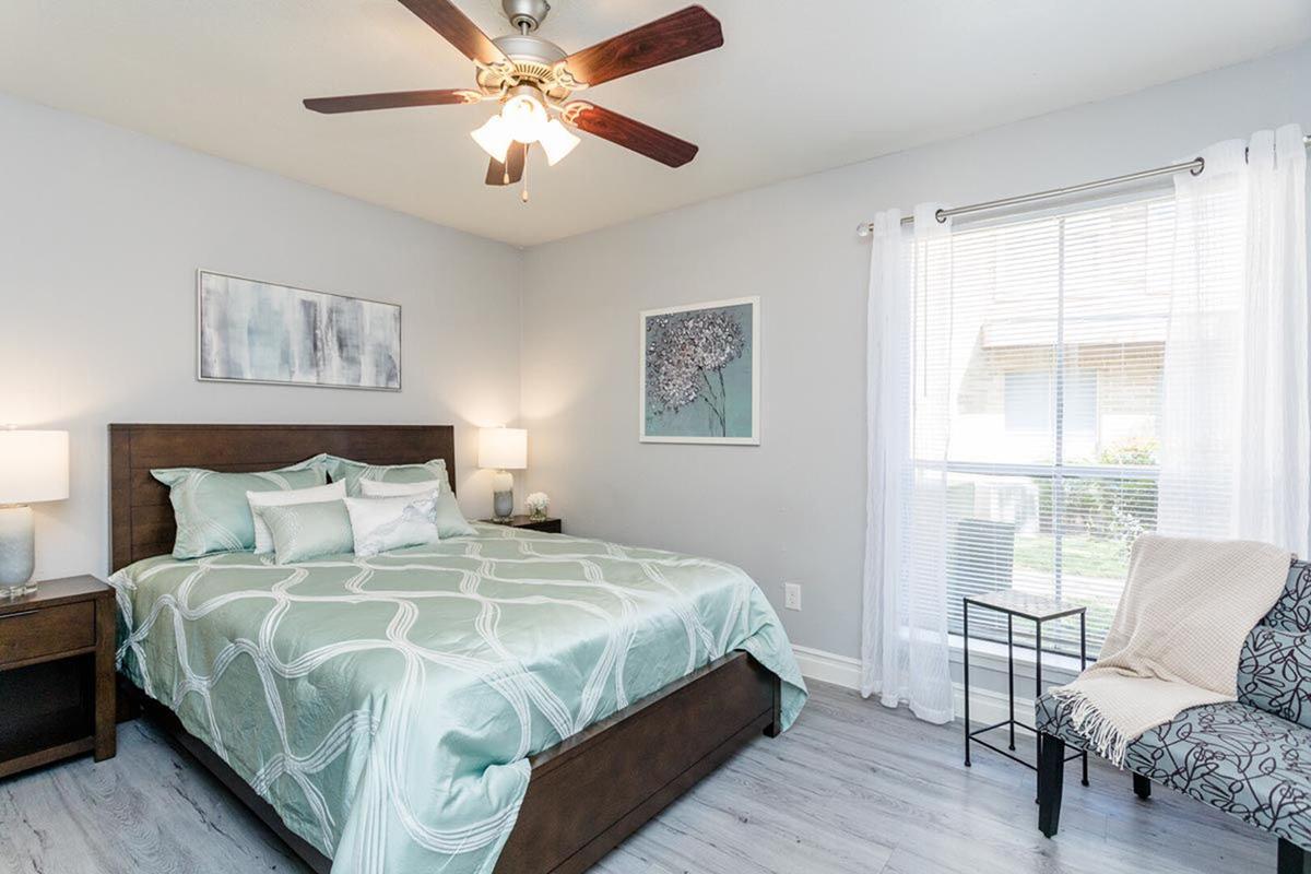 A well-decorated bedroom featuring a queen-sized bed with a light green patterned duvet, two bedside lamps, a ceiling fan, and a window with sheer white curtains. Additional decor includes two framed artworks on the walls and a small accent chair beside the window. The floor is a light-colored laminate.