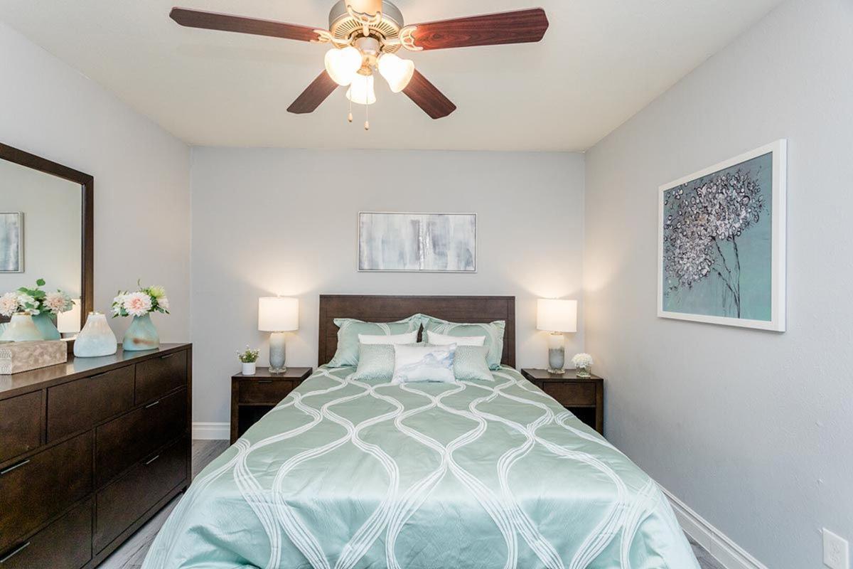A cozy bedroom featuring a wooden bed with a mint green quilt, two bedside lamps, and a dark wooden dresser. The walls are light gray, with a framed artwork hanging above the bed. A ceiling fan with three lights adds warmth to the space, complemented by decorative flowers on the dresser.