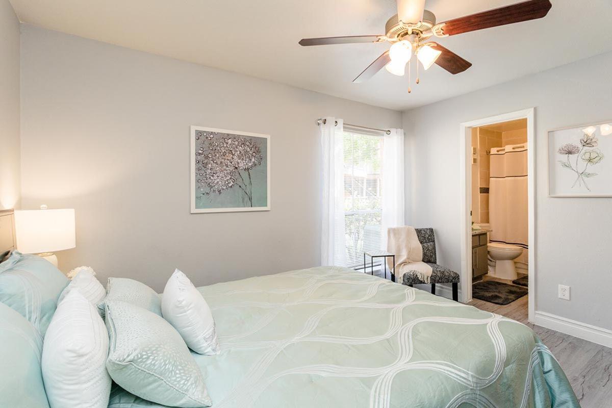 A cozy bedroom featuring a queen-sized bed with a light blue quilt, soft pillows, and a ceiling fan. Bright natural light enters through a window. There are decorative art pieces on the walls, and a door leads to a bathroom area. A chair is placed next to the bed, adding to the inviting atmosphere.