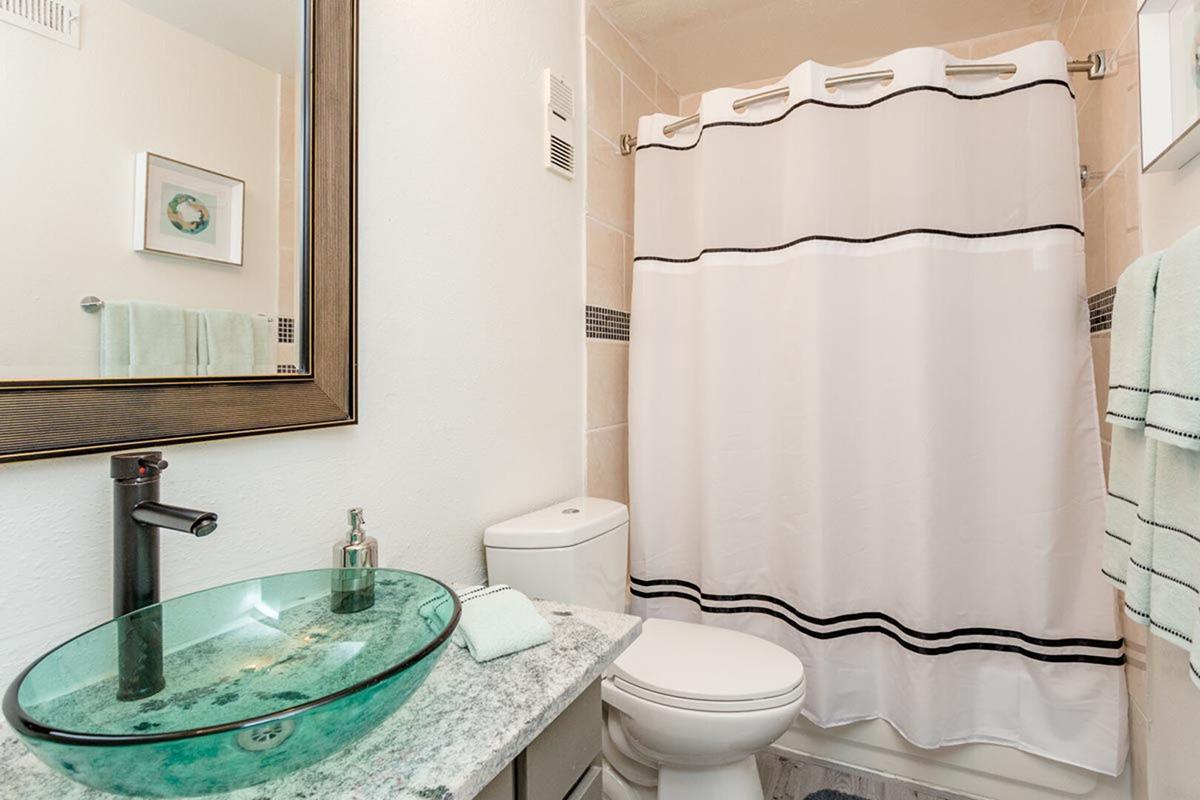 A modern bathroom featuring a white shower curtain with black stripes, a glass vessel sink on a granite countertop, a sleek black faucet, and a white toilet. Light-colored walls and a mirror enhance the space, complemented by a light green towel on the countertop.