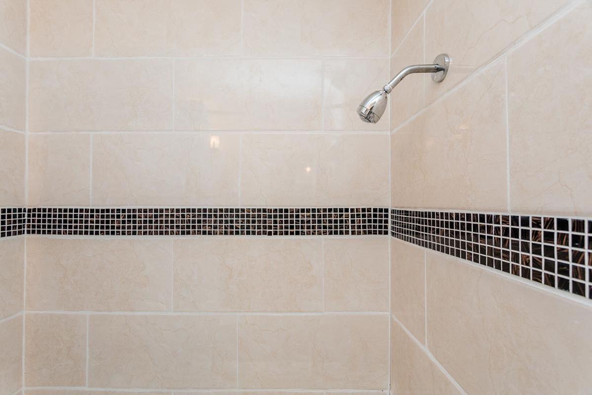 A close-up view of a tiled shower wall featuring light-colored tiles with a horizontal black mosaic stripe. The showerhead is mounted on the wall, and the overall appearance is clean and modern, showcasing a minimalist design.