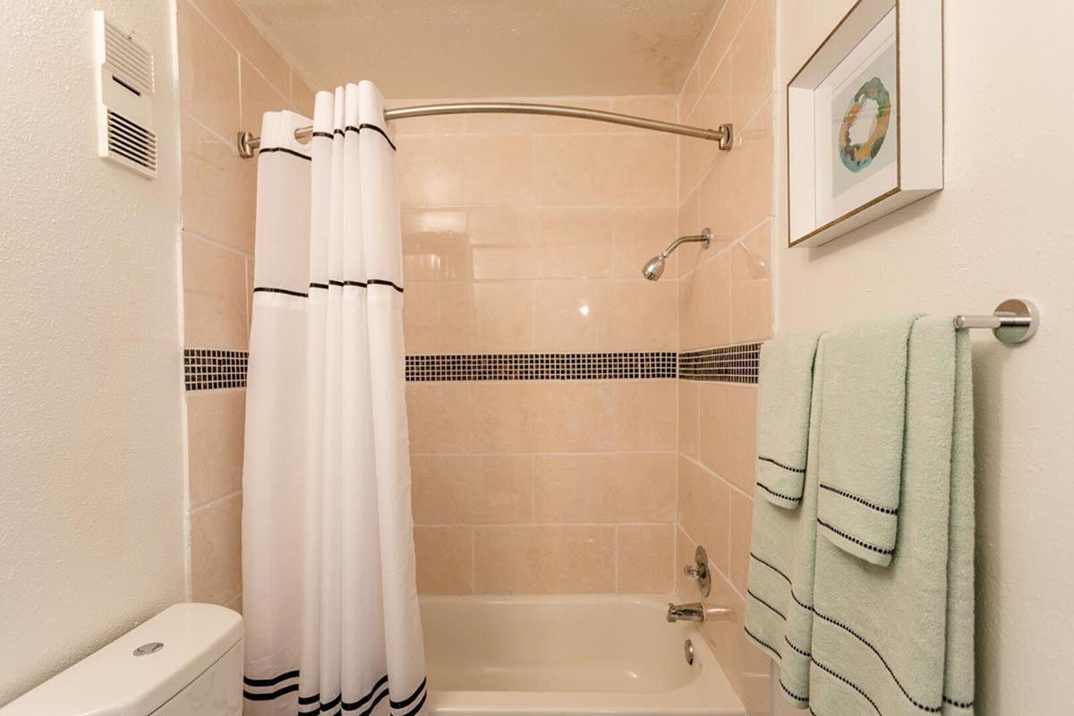 A clean bathroom featuring a bathtub with a shower curtain, a wall-mounted showerhead, and a towel rack with a light green towel. The walls are tiled with light-colored tiles, and there's a framed artwork hanging near the shower, adding a decorative touch.