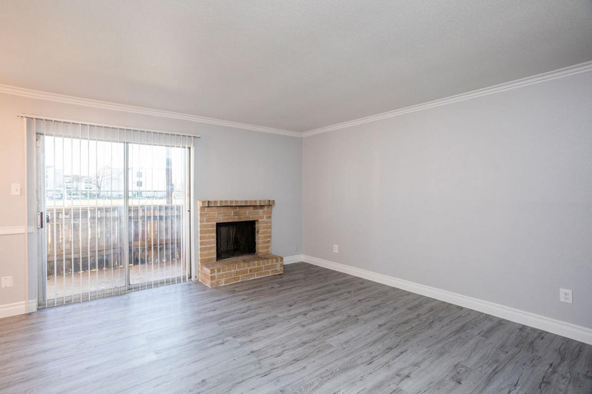 A spacious living room with light gray walls and wood-like flooring. There is a brick fireplace on one side and large sliding glass doors that lead to a balcony. Natural light enters the room, creating a bright and open atmosphere. The space is unfurnished, allowing for customizable decor.