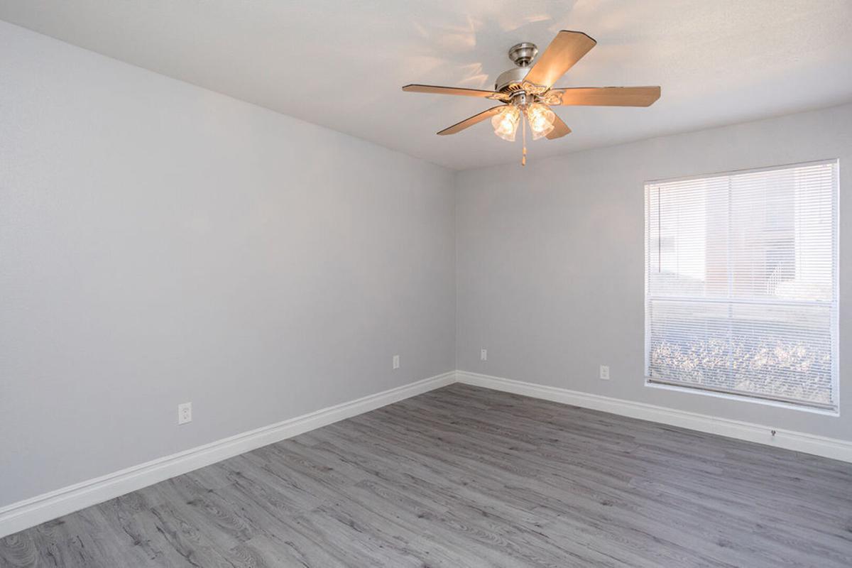 A clean and empty room featuring light gray walls, a ceiling fan with four light bulbs, and a large window with blinds. The flooring is light wood laminate, creating a bright and spacious atmosphere.
