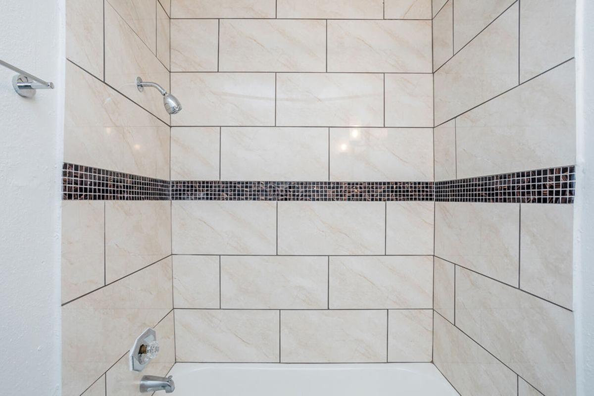 A clean and modern shower enclosure featuring light-colored tiled walls with a contrasting dark mosaic tile accent band. The showerhead is mounted on the wall, and a bathtub is visible below. The overall design is sleek and stylish, ideal for a contemporary bathroom setting.