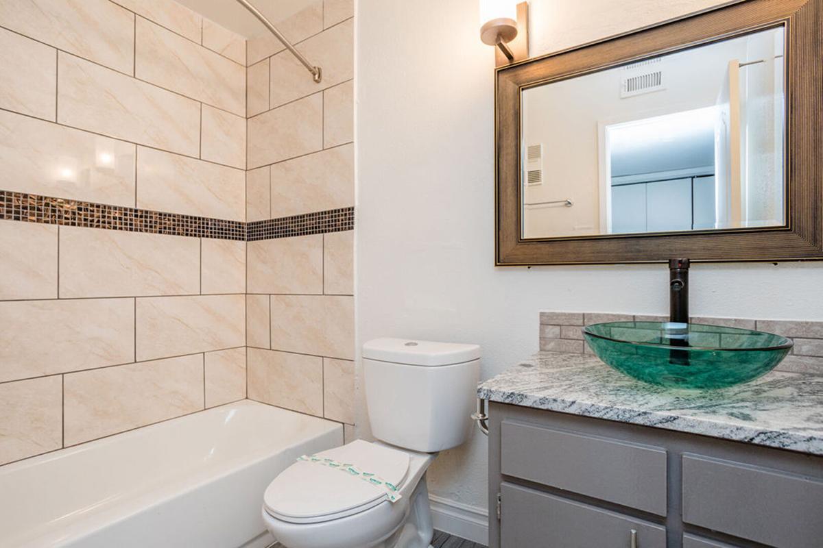 A modern bathroom featuring beige tiled walls, a bathtub, and a toilet. A granite-top vanity with a green glass sink and a large framed mirror is visible. Soft lighting is provided by a wall-mounted fixture above the mirror, adding warmth to the space.