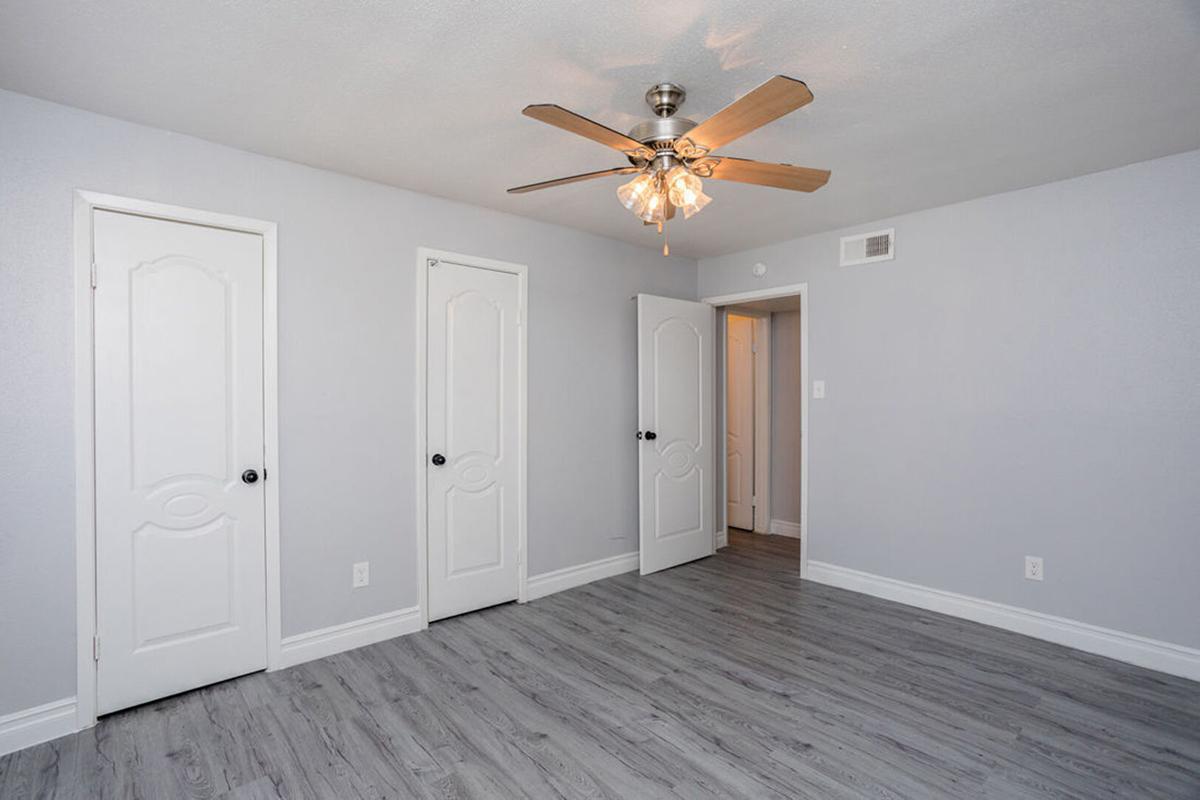 A spacious, well-lit bedroom featuring light gray walls and wood-style flooring. Three doors are visible: two closed and one slightly ajar, leading to another area. A ceiling fan with light fixtures hangs in the center, adding to the room's modern look. The atmosphere is clean and inviting.