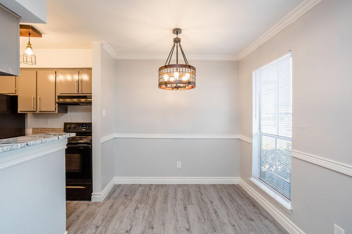 Modern kitchen area featuring a stylish chandelier, gray walls, and hardwood-like flooring. There are dark cabinets and an oven visible, along with a window allowing natural light to enter. The space is well-lit and contemporary in design, creating an inviting atmosphere.