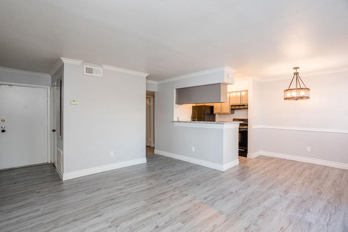 Bright and spacious living area featuring light gray walls and a modern laminate floor. A small kitchen area with dark cabinets is visible in the background, along with a stylish pendant light hanging from the ceiling. The room is well-lit, creating a welcoming atmosphere. An entry door leads to the left.