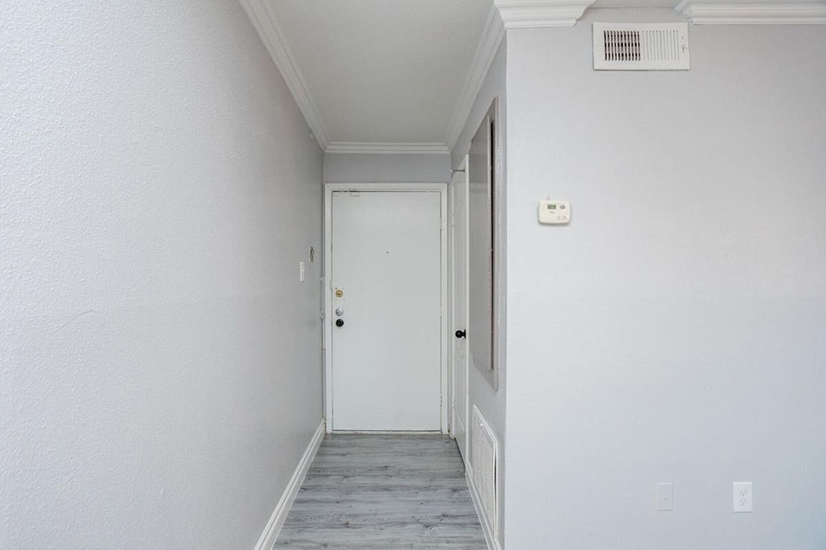 A narrow hallway with light gray walls and a white door at the end. The floor is a light-colored laminate, and there is a visible wall-mounted thermostat on the right side. The ceiling features a simple crown molding, and there is a small, framed mirror on the left wall.