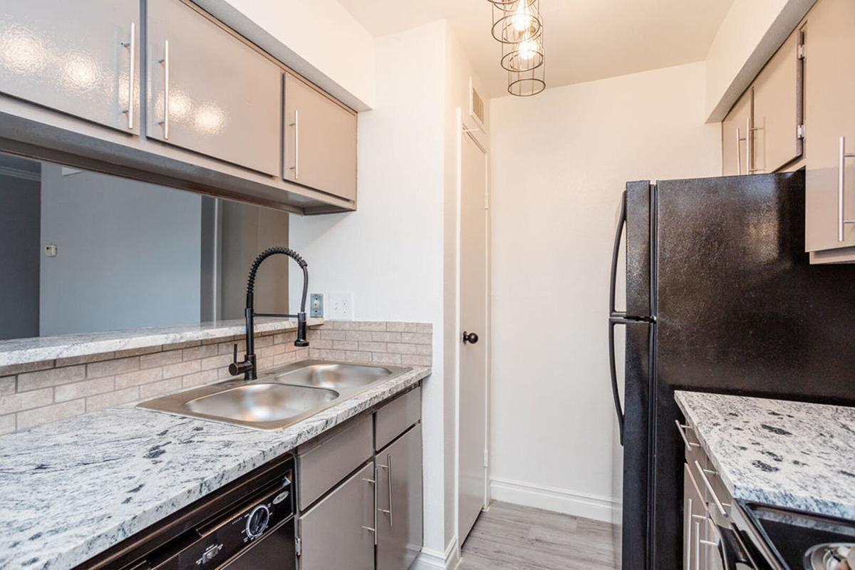 Modern kitchen featuring gray cabinetry, granite countertops, double sink, and a black refrigerator. Located in a well-lit space with pendant lighting hanging above. The walls are painted white, and there is a small adjacent hallway or doorway visible in the background.