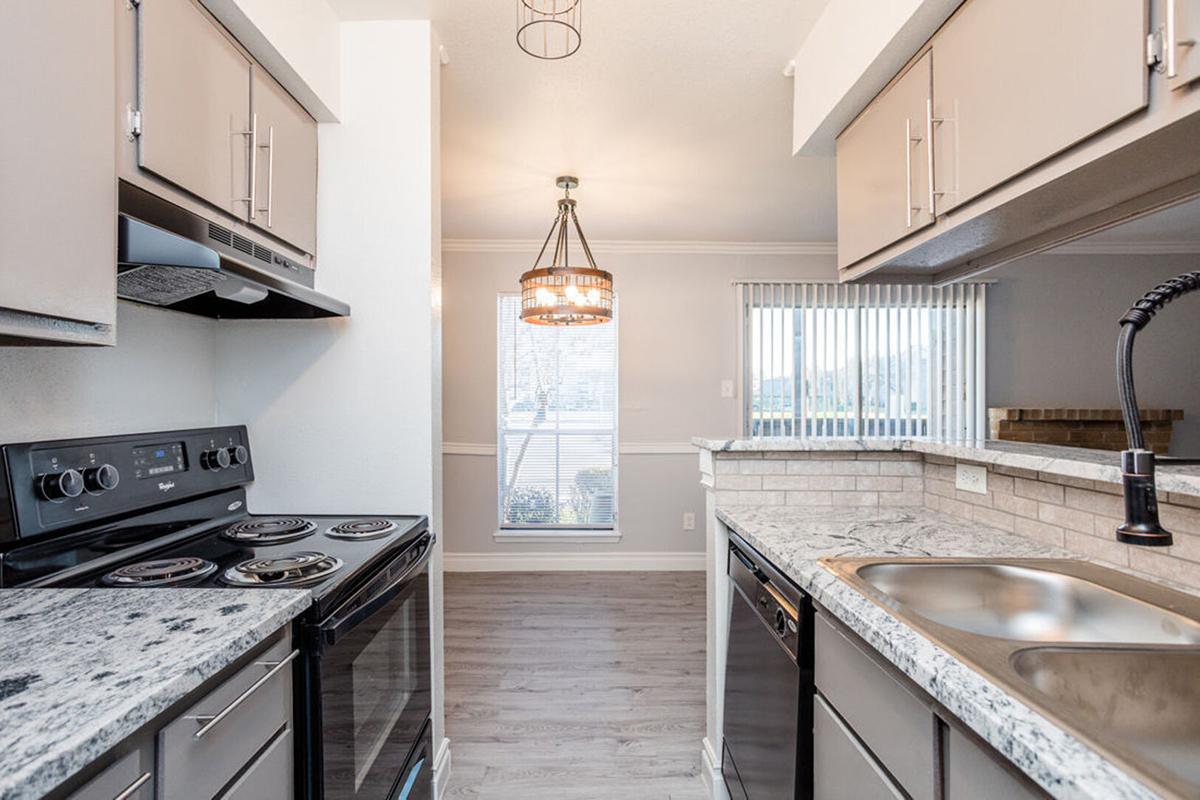 Modern kitchen featuring dark and light cabinetry, granite countertops, a stainless steel sink, and a black stove. A pendant light hangs above the dining area, which is illuminated by natural light coming through a window with white blinds. The floor is made of light wood laminate.