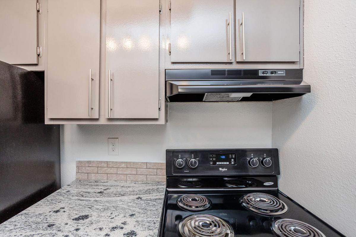 A modern kitchen featuring a black stove with four burners, an overhead range hood, and light-colored cabinets. The countertops are made of granite with a subtle pattern, and there's a small tile backsplash. The overall layout appears clean and well-maintained.