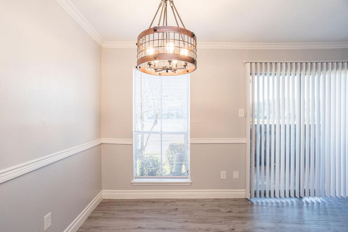 A well-lit room featuring a modern chandelier hanging from the ceiling. There is a large window with vertical blinds allowing natural light to enter. The walls are painted in a light color, and the flooring is a sleek, smooth wood finish. A cozy and inviting atmosphere is created in this space.