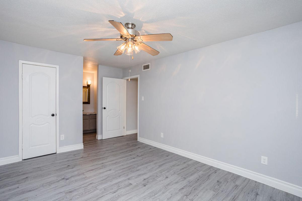A spacious room with light gray walls and wooden flooring. A ceiling fan with light fixtures is centered above. There are two doors: one leads to another room and the other is probably a closet. A bathroom is visible in the background, shown by a light reflection. The overall ambiance is bright and clean.