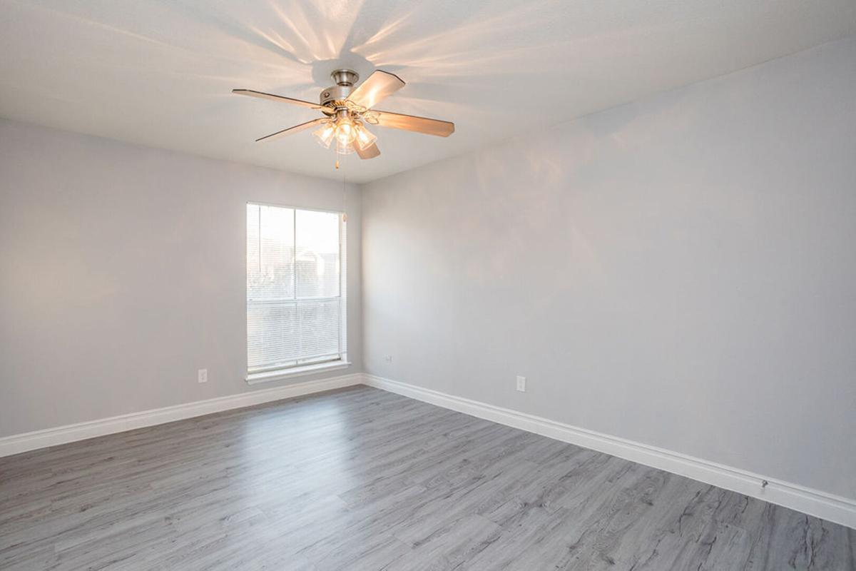 A spacious, empty room with light gray walls and laminate flooring. A ceiling fan hangs from the ceiling, and a window lets in natural light, creating a bright atmosphere. The room is devoid of furniture, emphasizing its open layout.