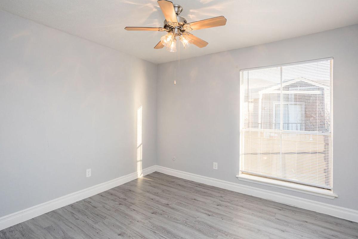 A well-lit, empty room featuring light gray walls, a modern ceiling fan with wooden blades, and a window with blinds allowing natural light to enter. The floor is a light-colored laminate that enhances the space's airy feel. Ideal for a fresh start or as a blank canvas for interior design.