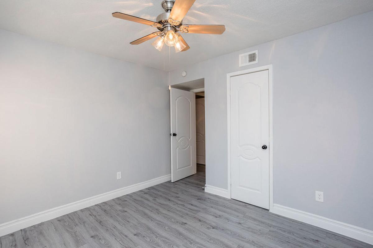 Empty room with light gray walls and a light wooden floor. A ceiling fan with five blades is mounted in the center. There is a doorway leading to another space, and a closed white door on the left side of the image. The overall atmosphere is bright and minimalist.
