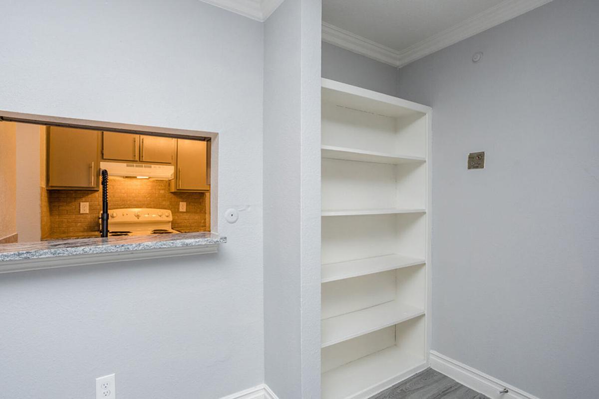 A light gray room featuring a small kitchen pass-through with an oven visible. To the right, there is a white shelf unit built against the wall. The overall design is modern and minimalistic, with neutral colors and clean lines.