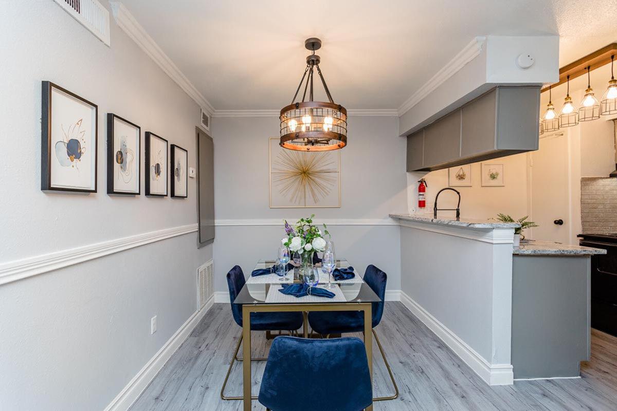 A modern dining area featuring a table set for dinner with navy blue chairs, elegant tableware, and a floral centerpiece. Wall-mounted art pieces enhance the space, and a stylish light fixture hangs above the table. An open kitchen area is visible in the background, with pendant lights illuminating the counter.
