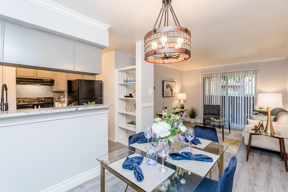 A modern dining area featuring a glass table set with blue napkins and clear glassware. In the background, a cozy living space with a beige sofa, decorative plants, and a sliding glass door. A stylish pendant light hangs above the dining table, and the kitchen can be seen through an open layout.