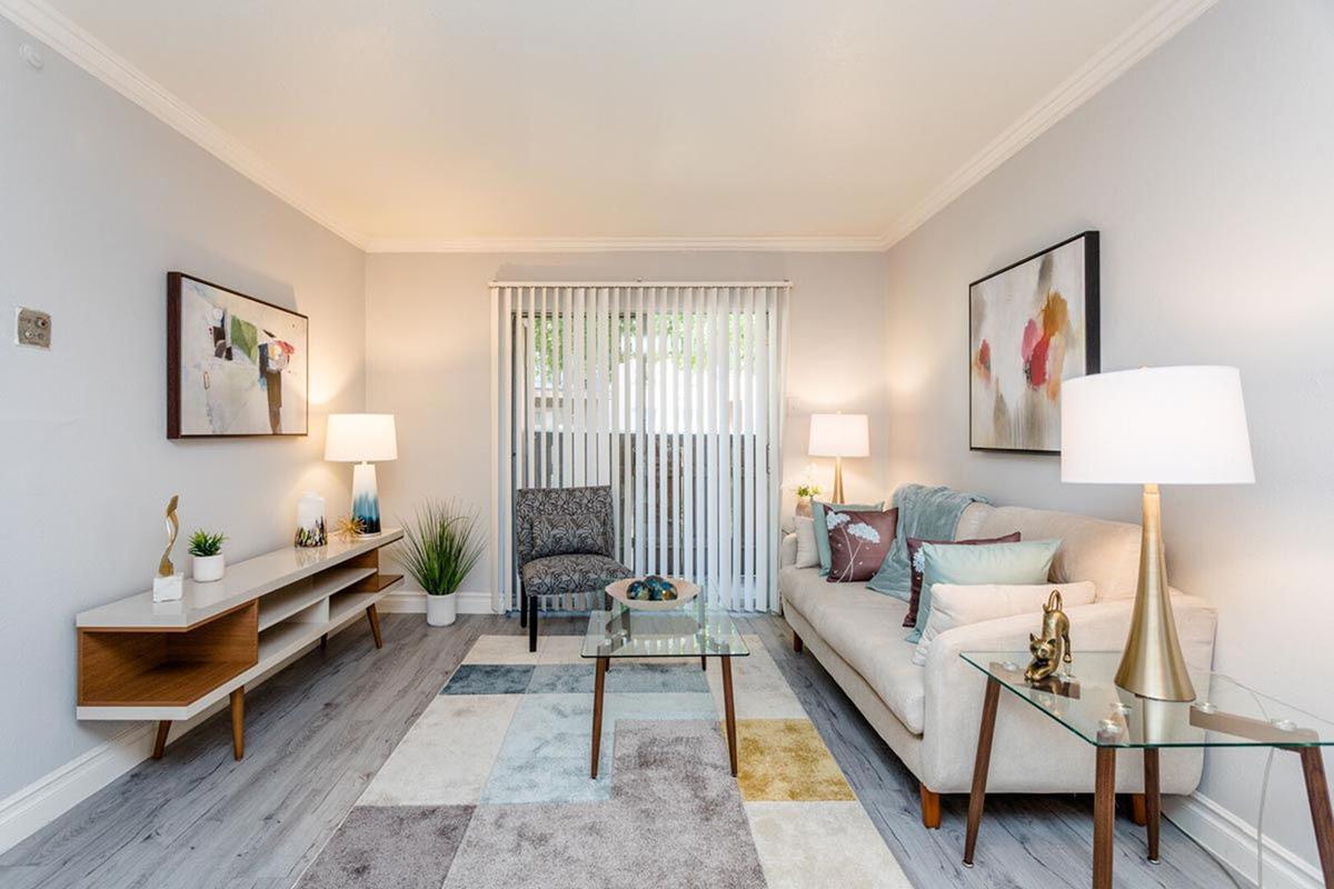 A bright and airy living room featuring a light-colored sofa with decorative pillows, a patterned area rug, two stylish lamps, and a modern wooden console table. There is a decorative plant, a chair in the corner, and sheer blinds allowing natural light to fill the space, complemented by tasteful artwork on the walls.