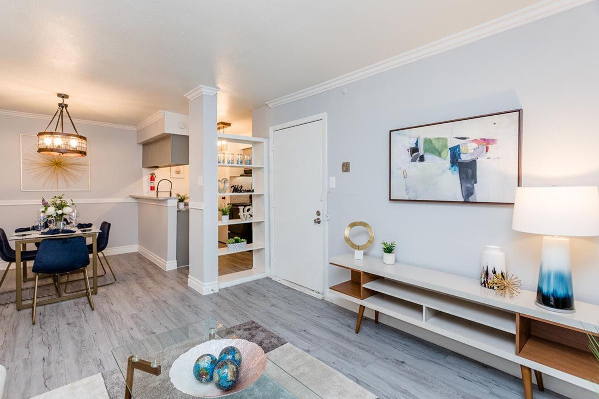 Modern living room featuring a light gray color scheme, a stylish coffee table with decorative items, a console with an abstract painting above, and a cozy dining area with a round table and blue chairs in the background. There’s a soft glow from a hanging light fixture and a welcoming atmosphere.