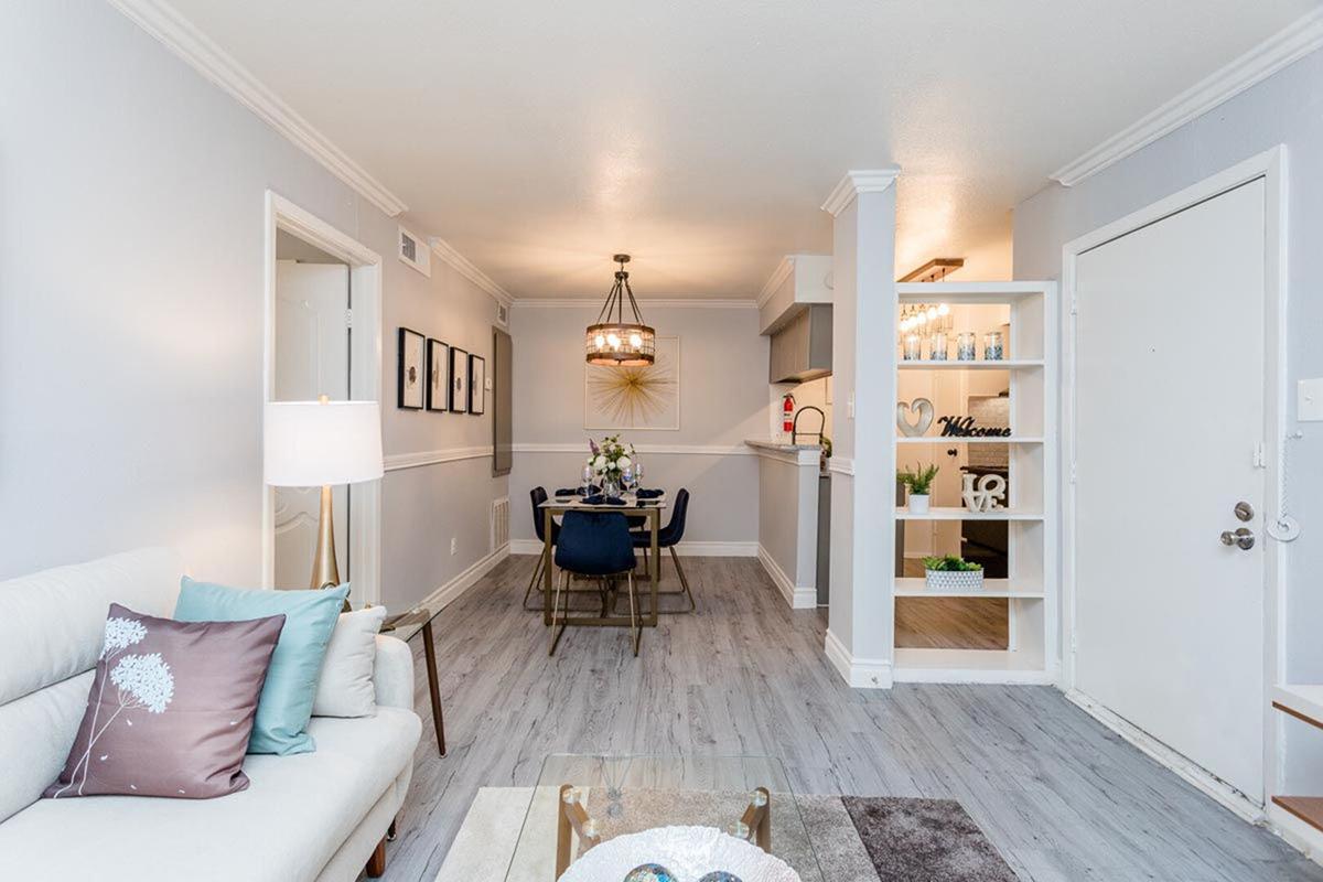 A cozy, modern living space featuring a light gray wall color, a comfortable beige sofa with decorative pillows, a dining area with a dark wooden table and chairs, and a stylish chandelier. A shelf displays decorative items, while a welcoming atmosphere is created by soft lighting and tasteful decor.