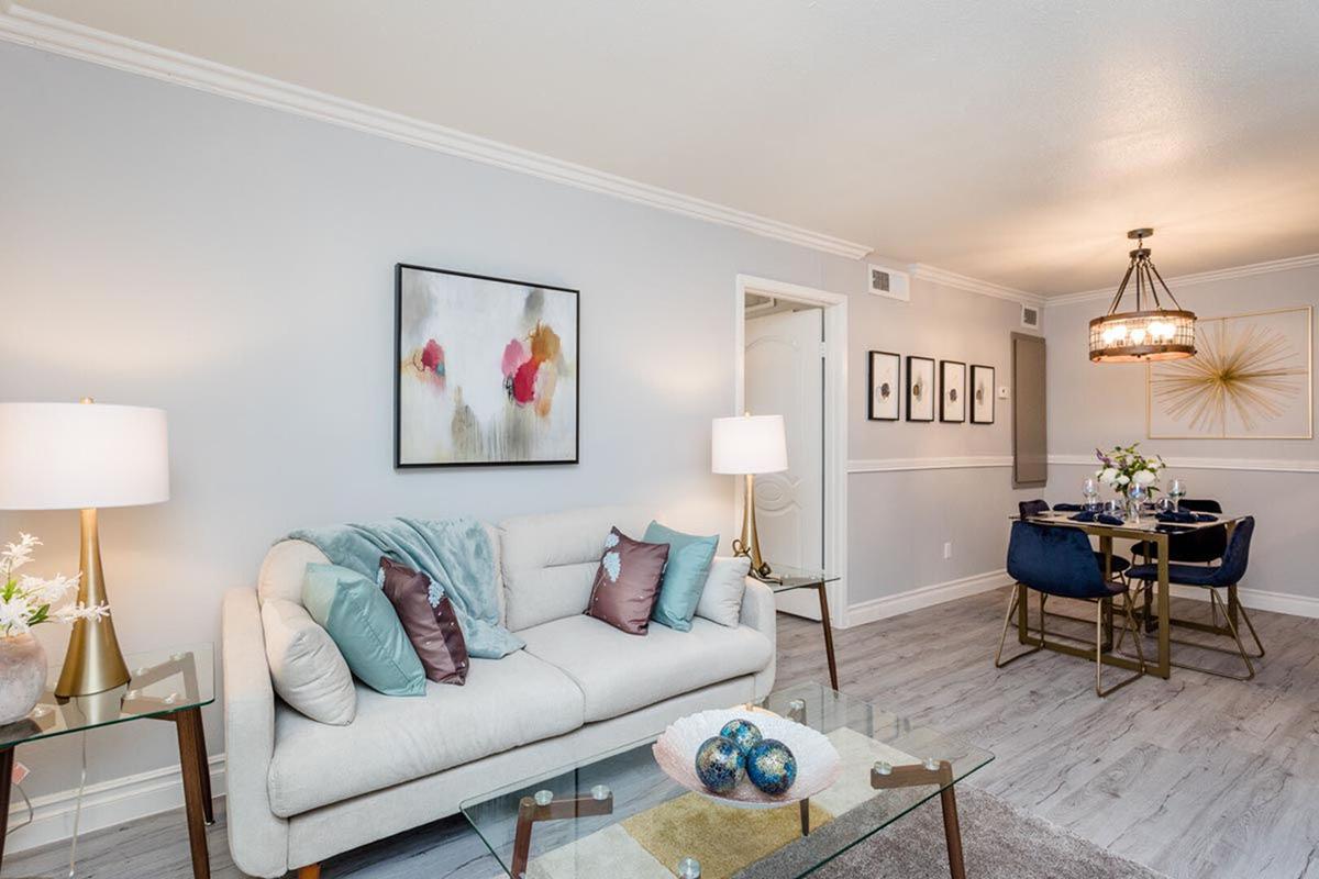 A stylish living room featuring a light-colored sofa with decorative pillows, a glass coffee table, and a piece of abstract art on the wall. To the right, a dining area with a round table, blue chairs, and a floral centerpiece, all set against a light gray wall and warm lighting.