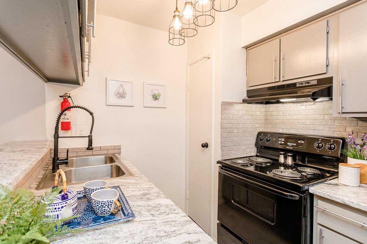 A modern kitchen featuring gray cabinetry, a black stove, and a silver hood. The countertop is made of light-colored stone, and there are two sinks. Decorative items include a plant, blue and white dishware, and framed art on the wall. A stylish light fixture hangs from the ceiling, adding ambiance.