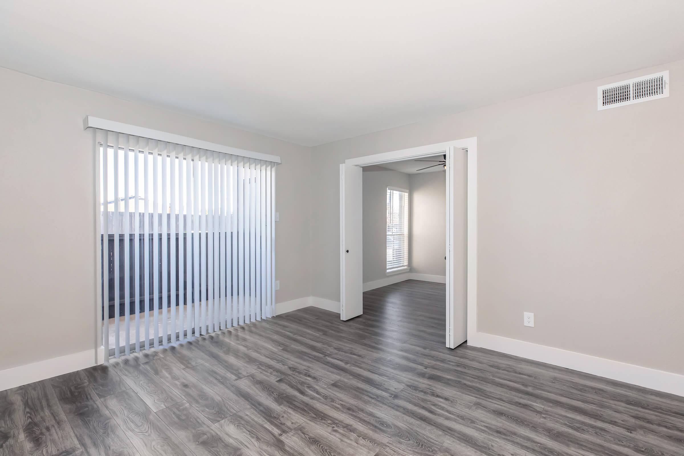A modern, empty room featuring light-colored walls and vinyl flooring. To the left, there are vertical blinds covering a sliding glass door leading to a balcony. An open doorway leads to another room visible in the background. The setup creates a bright and spacious atmosphere.