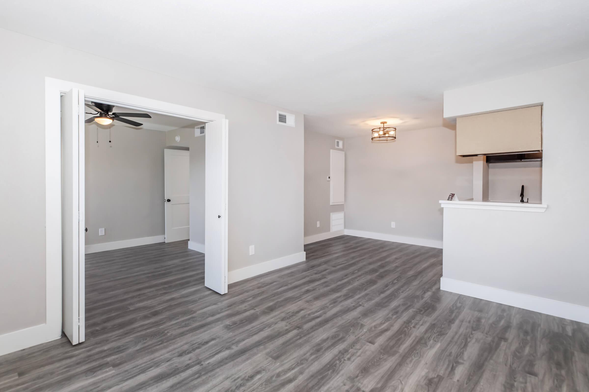 Bright, spacious living area featuring light gray walls and modern laminate flooring. A doorway leads to another room, while a small kitchen space with a bar counter is visible in the background. A ceiling fan adds to the comfort of the space. Natural light fills the area, creating a welcoming atmosphere.