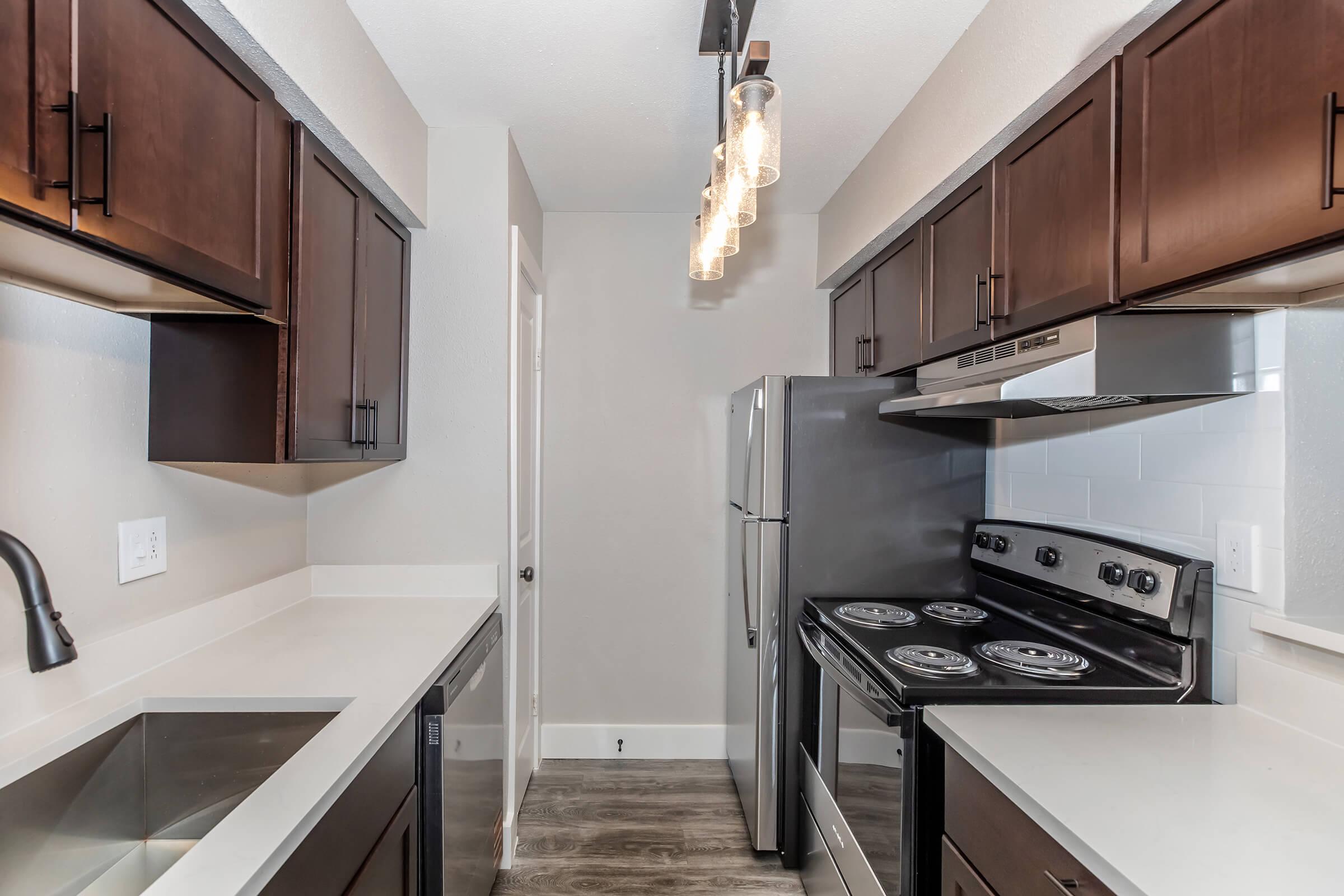 Modern kitchen featuring dark wooden cabinetry, sleek countertops, stainless steel appliances including a refrigerator and oven, and a stylish pendant light. The space is well-lit with neutral-colored walls, showcasing an open layout and contemporary design elements.