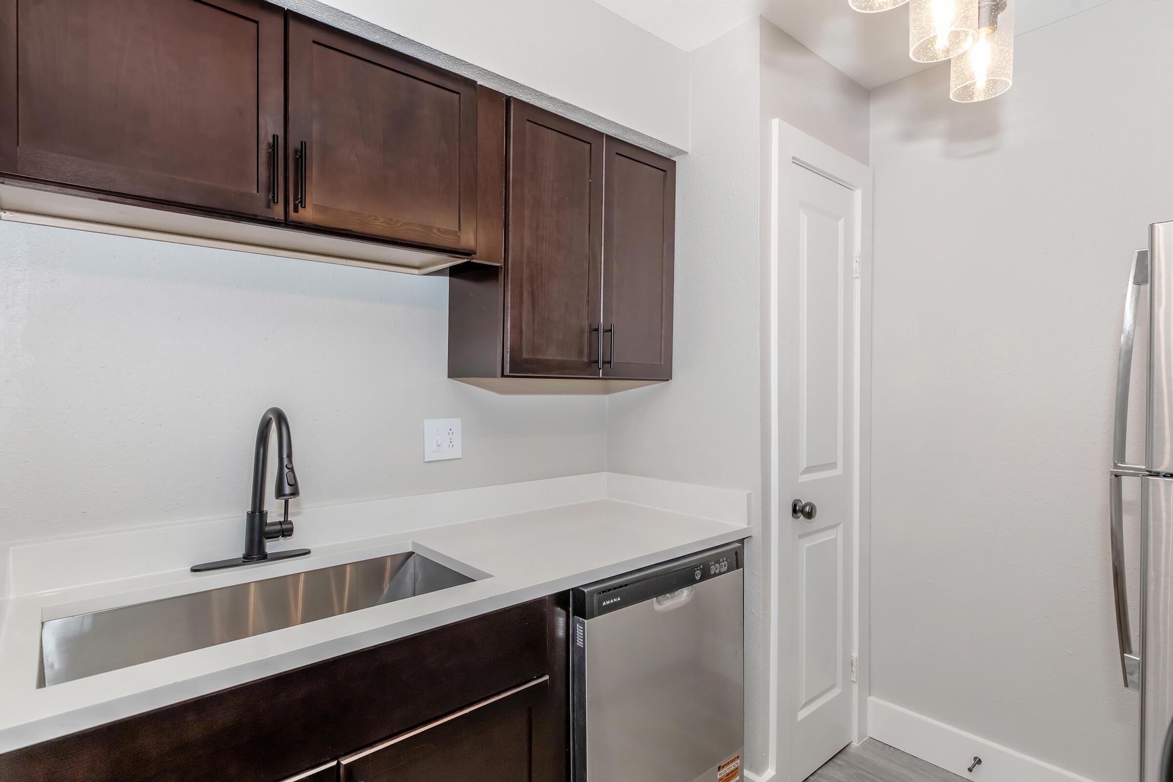 Modern kitchen featuring dark wood cabinets, a stainless steel sink with a sleek black faucet, and a silver dishwasher. A stainless steel refrigerator is visible, and pendant lights hang above the countertop. The walls are a light gray, creating a bright and contemporary look.