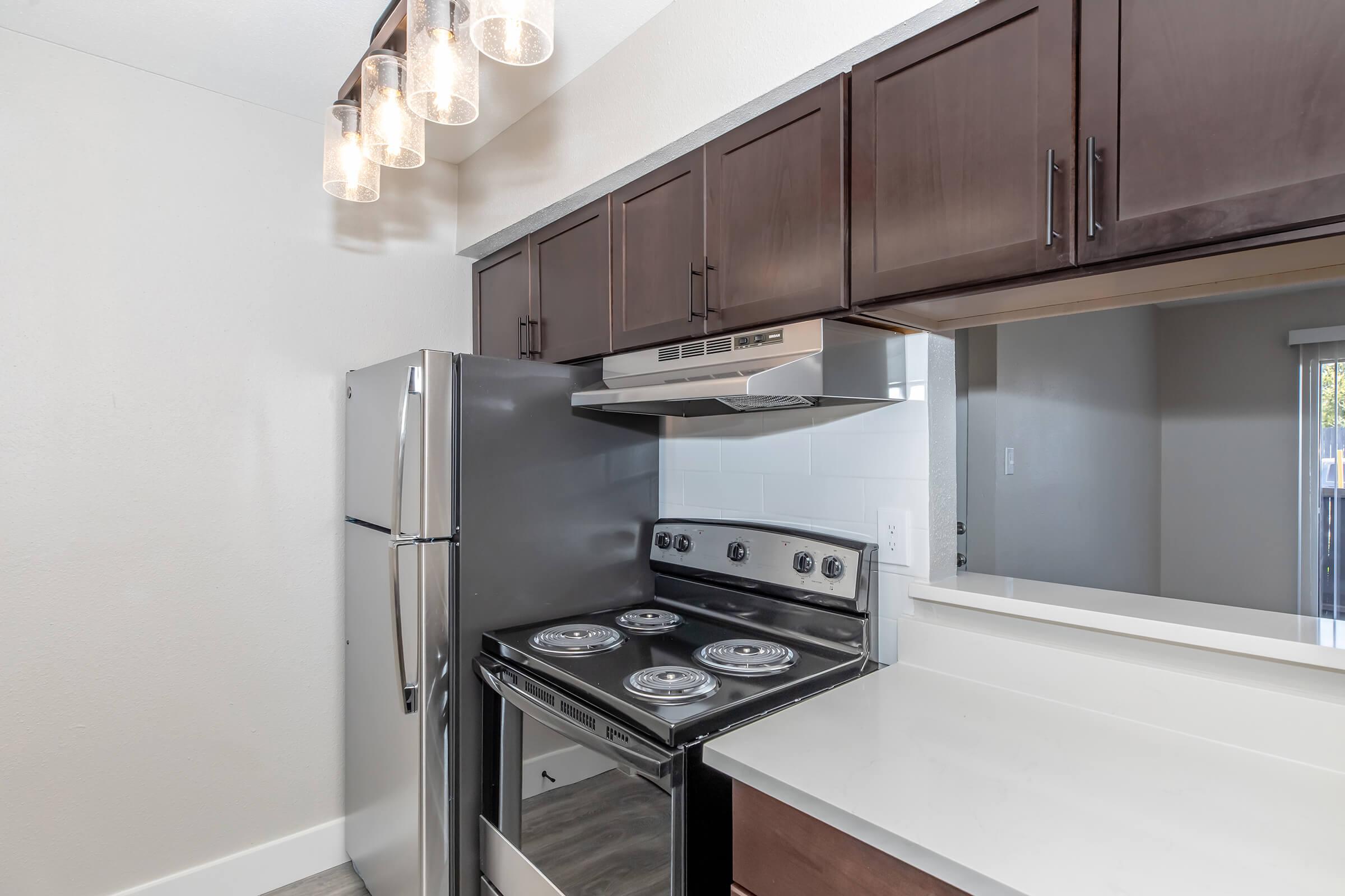 Modern kitchen featuring dark wood cabinetry, stainless steel refrigerator, and a black stove with four burners. The countertop is light-colored, and there are pendant lights above the kitchen island, providing bright overhead illumination. A window is visible in the background.