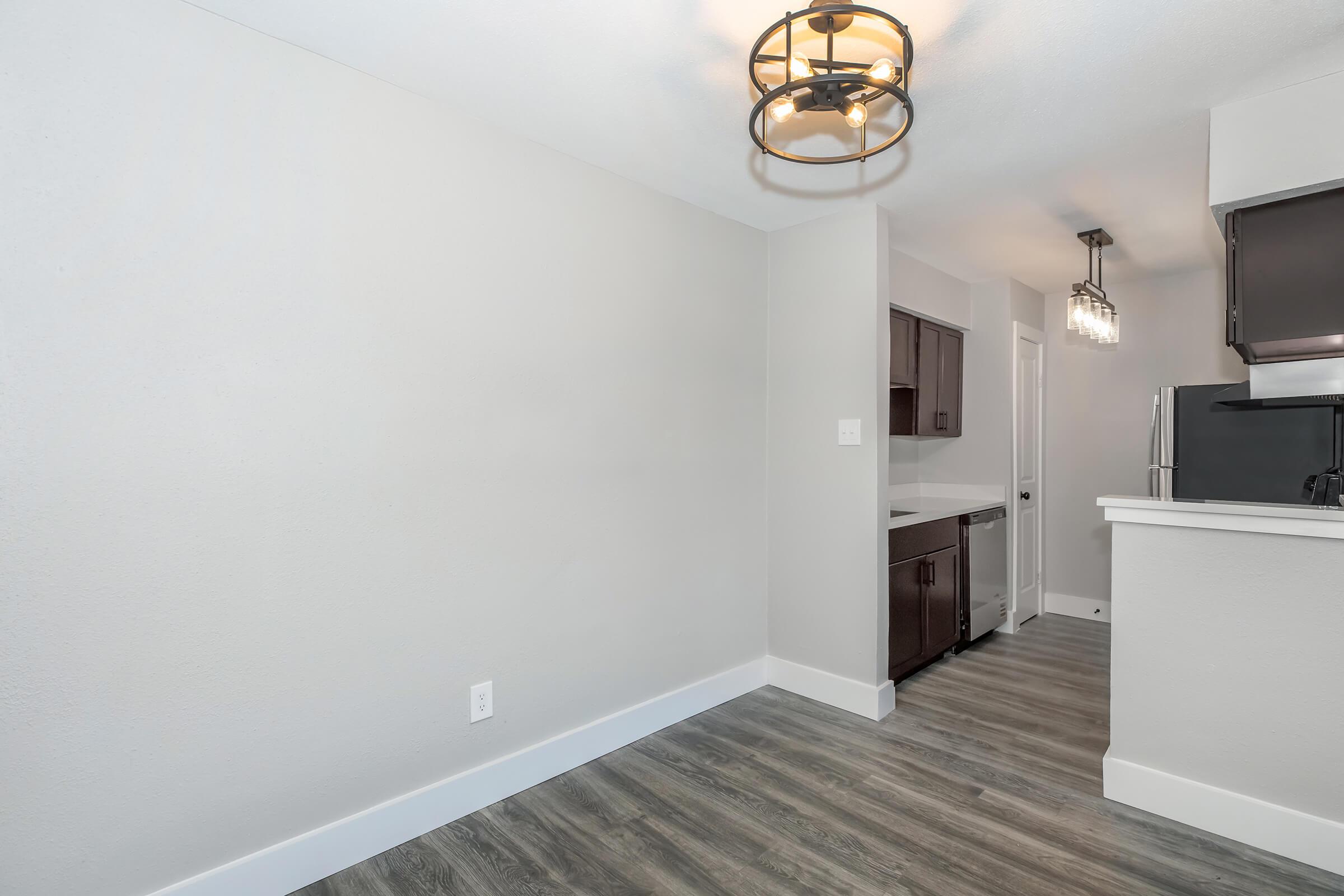A modern kitchen with gray walls, sleek dark cabinetry, and stainless steel appliances. The room features a hardwood floor, a bright ceiling light fixture, and an open layout that connects to a dining area. The space is well-lit and has a clean, contemporary design.