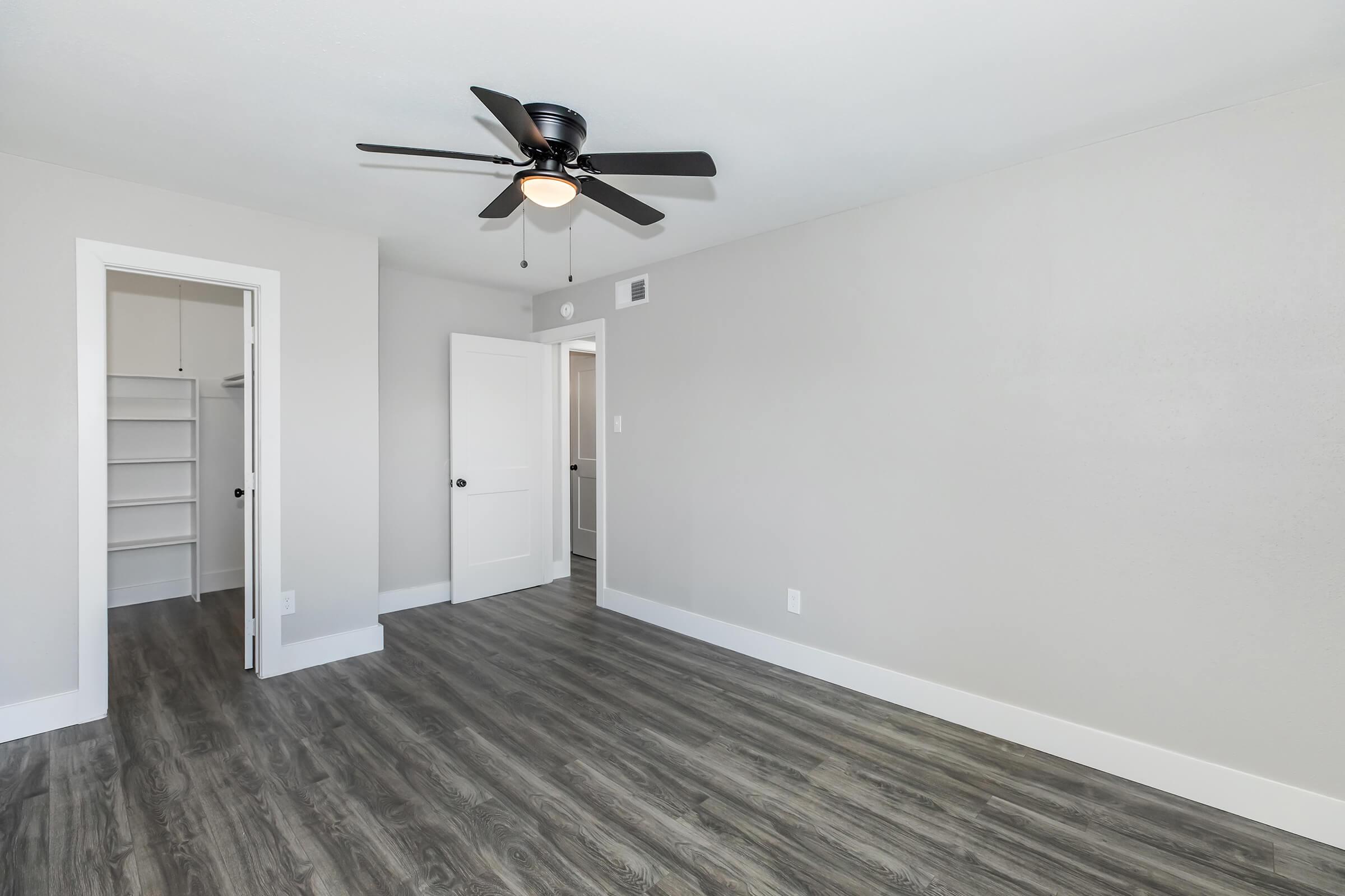 Empty room featuring a ceiling fan, light-colored walls, and hardwood-style flooring. A door leads to a closet on one side, while another door opens to a hallway. The space is well-lit and has a modern, minimalist design.