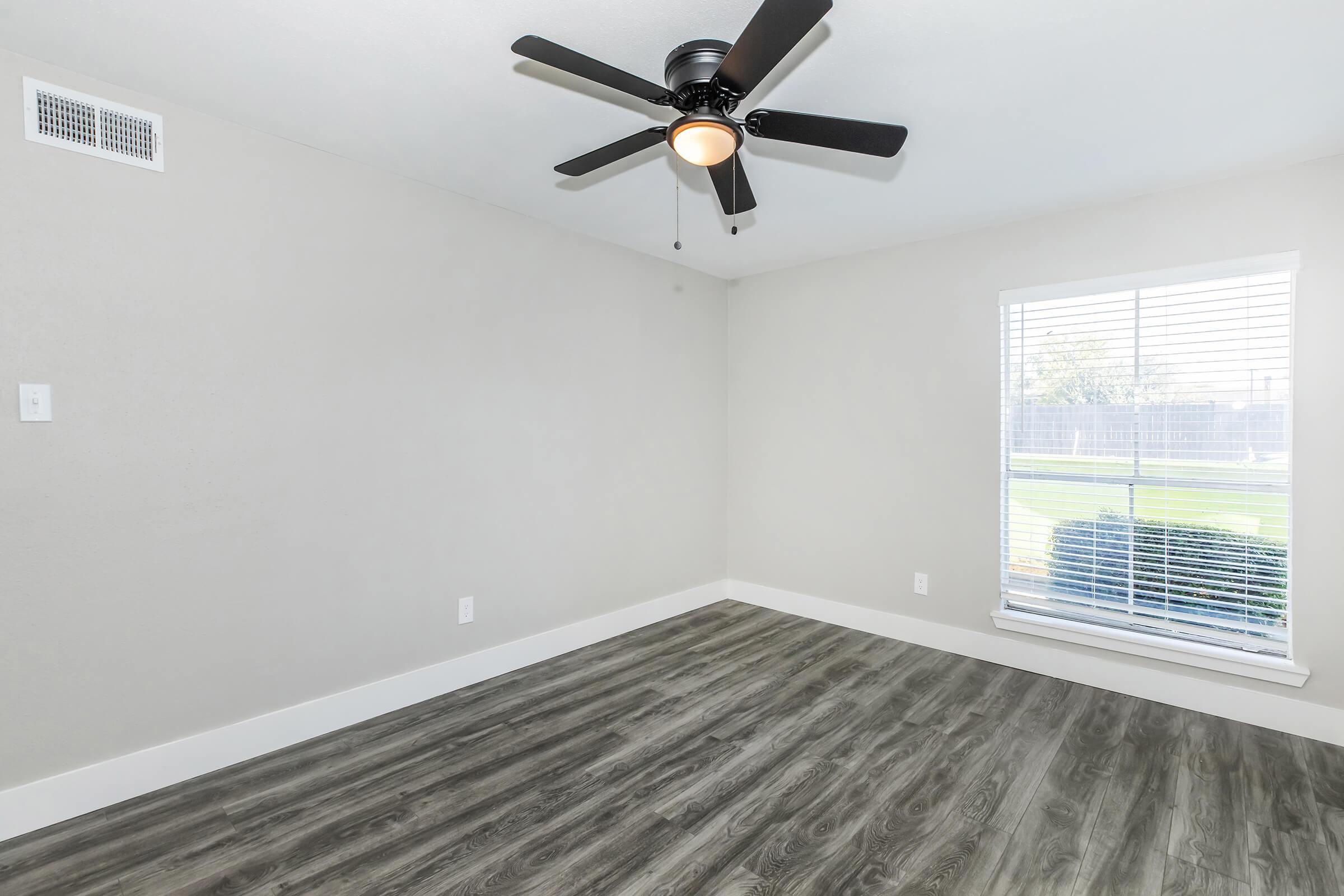 Empty room with light gray walls and a dark ceiling fan. The floor features wood-like laminate in a muted tone. A large window with white blinds allows natural light to enter, revealing a view of greenery outside. The room has a spacious and clean appearance, ideal for various uses.
