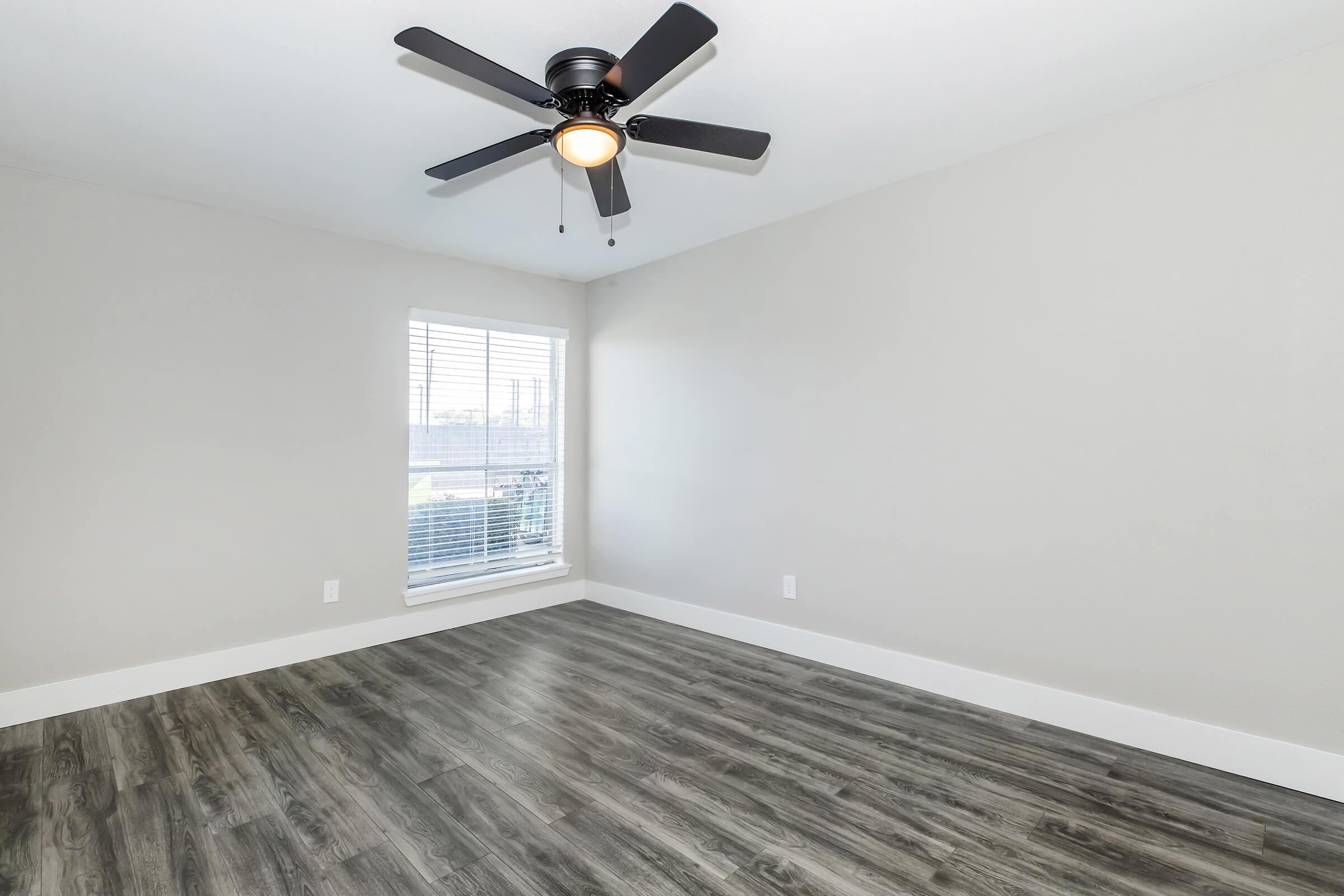 A bright, empty room with light gray walls and a dark wood laminate floor. A ceiling fan with a light fixture is mounted in the center, and there's a large window with closed blinds on one side, allowing natural light to enter the space.