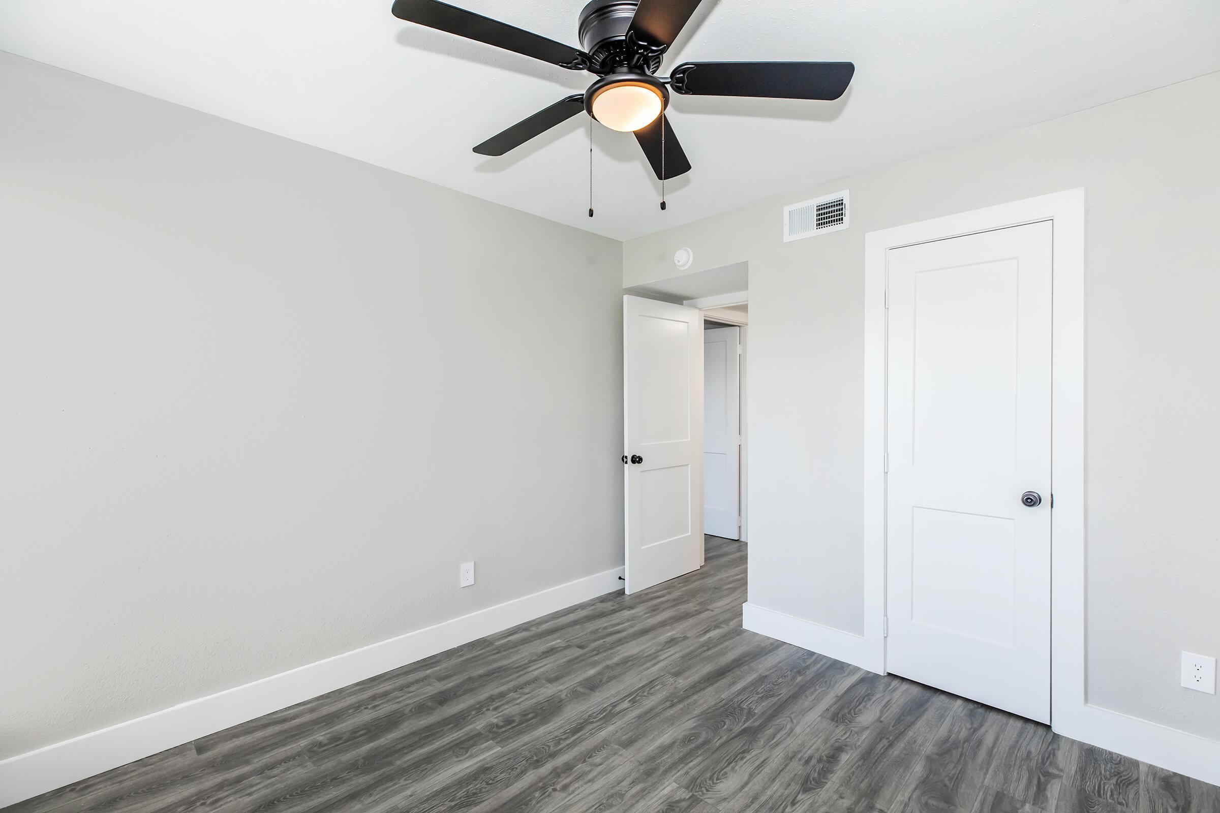 A modern, empty room with light gray walls and a ceiling fan. There is a door on the left leading to another room and a closet door on the right. The floor features wood-like laminate flooring, contributing to a sleek and clean aesthetic. The space is well-lit with natural light.